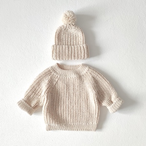 Puede incluir: Un su&eacute;ter de punto color crema y un gorro a juego con un pomp&oacute;n en la parte superior.