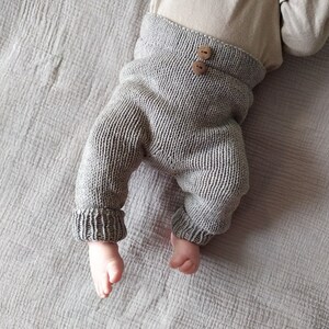 Op de afbeelding: Grijze gebreide babybroek met houten knopen. De broek is gemaakt van een zacht, comfortabel materiaal en heeft een ontspannen pasvorm.