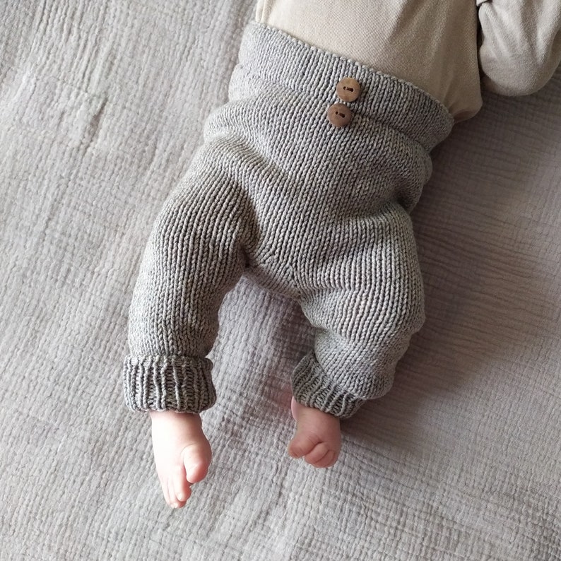 Op de afbeelding: Grijze gebreide babybroek met houten knopen. De broek is gemaakt van een zacht, comfortabel materiaal en heeft een ontspannen pasvorm.