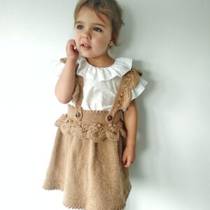 May include: A little girl wearing a brown knitted skirt with a white ruffled blouse. The skirt has a lace trim and a button closure.