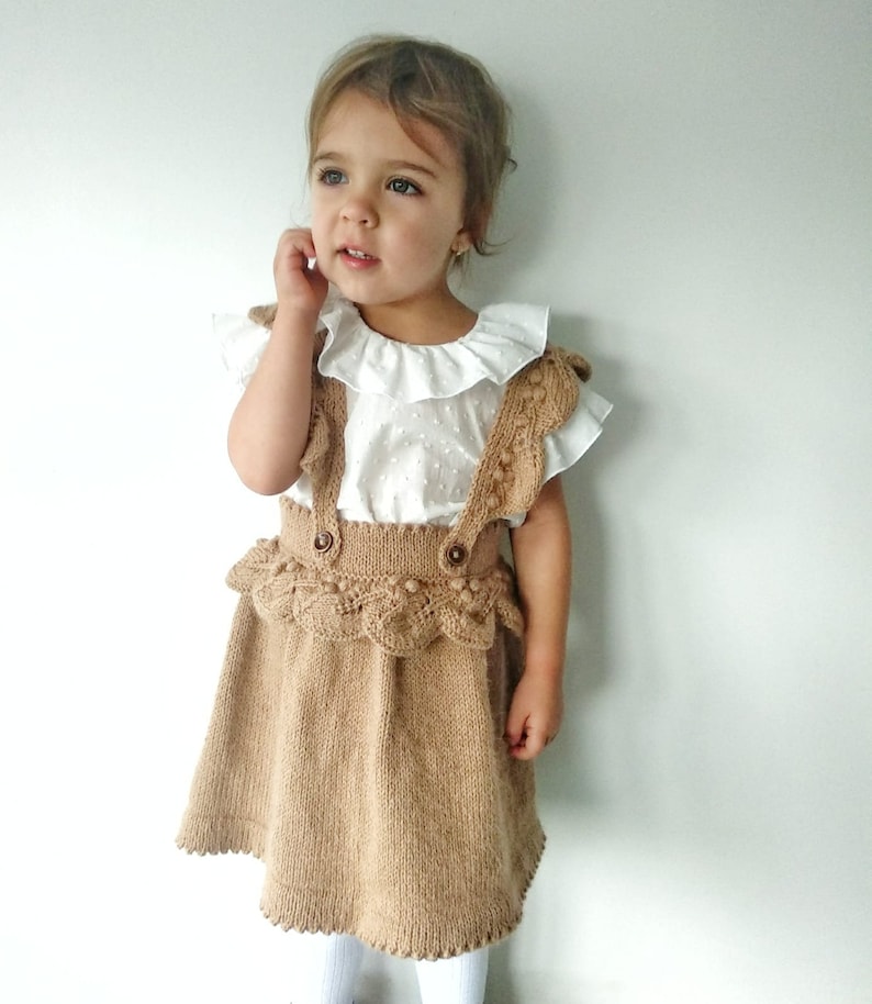 May include: A little girl wearing a brown knitted skirt with a white ruffled blouse. The skirt has a lace trim and a button closure.