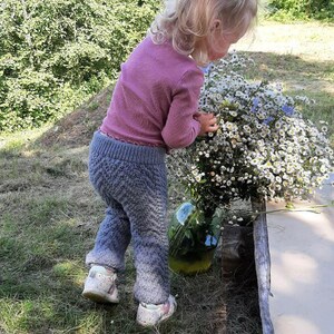 May include: A young girl wearing a pink long-sleeved shirt and grey knitted trousers is holding a bunch of white and blue wildflowers. She is standing in a grassy area with a wooden log and a glass jar filled with flowers in the background.