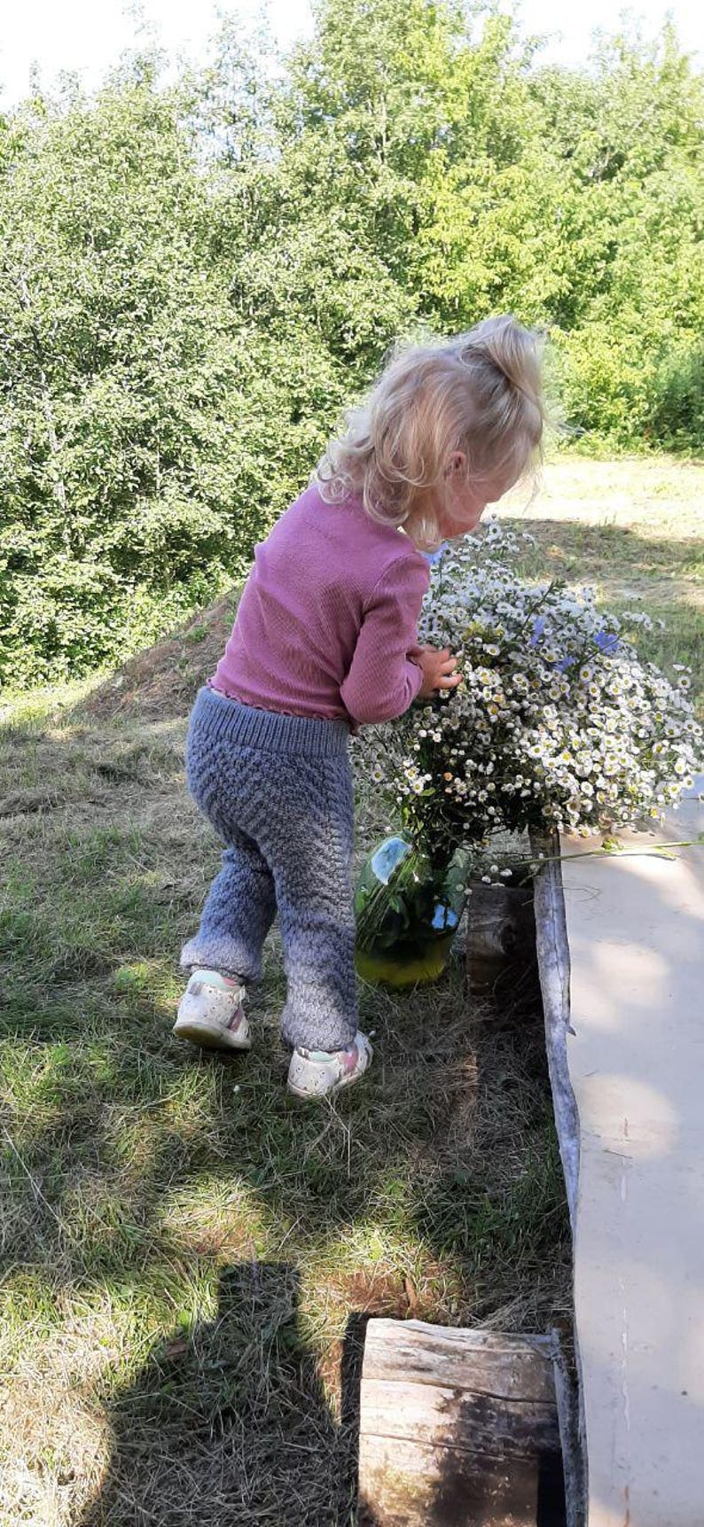 May include: A young girl wearing a pink long-sleeved shirt and grey knitted trousers is holding a bunch of white and blue wildflowers. She is standing in a grassy area with a wooden log and a glass jar filled with flowers in the background.