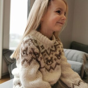 May include: A young girl wearing a cream-colored sweater with a brown geometric pattern. The sweater has long sleeves and a high neck. The girl is sitting on a couch and has long blonde hair.