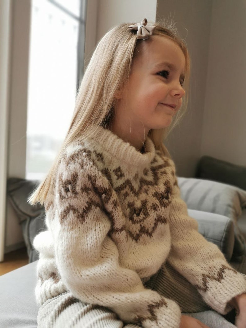 May include: A young girl wearing a cream-colored sweater with a brown geometric pattern. The sweater has long sleeves and a high neck. The girl is sitting on a couch and has long blonde hair.