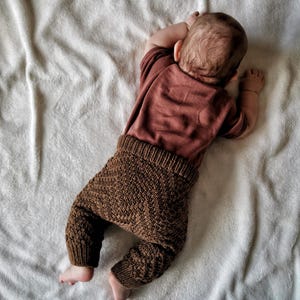 May include: A baby wearing brown knitted trousers and a maroon long-sleeved top, lying on a white fluffy blanket.