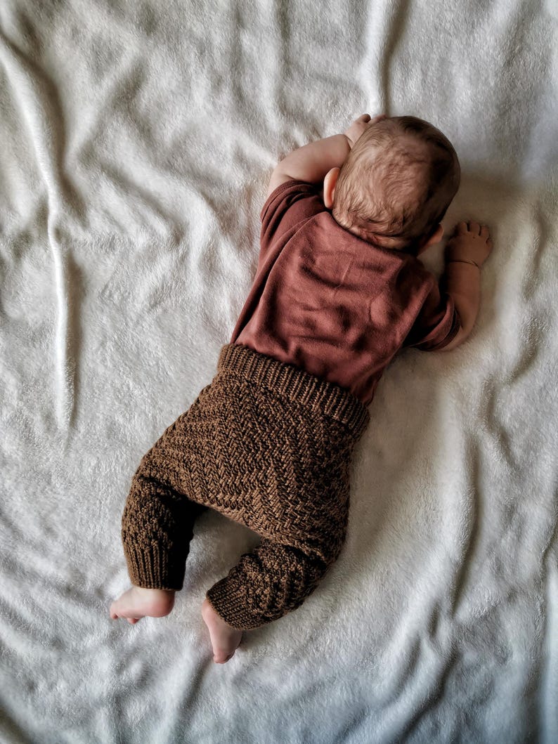 May include: A baby wearing brown knitted trousers and a maroon long-sleeved top, lying on a white fluffy blanket.