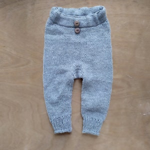 Op de afbeelding: Een lichtgrijze gebreide babybroek met twee houten knopen aan de voorkant.