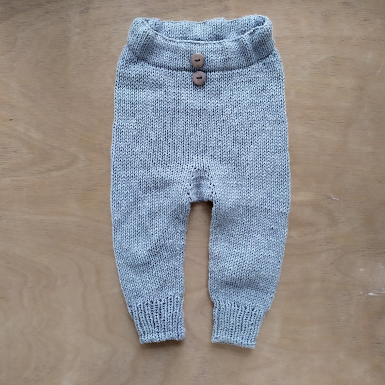 Op de afbeelding: Een lichtgrijze gebreide babybroek met twee houten knopen aan de voorkant.