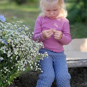 May include: A young child wearing a pink long-sleeved shirt and blue knitted trousers is sitting on a wooden surface. The child is holding a small white flower in their hand. The child is wearing white sandals with pink accents.