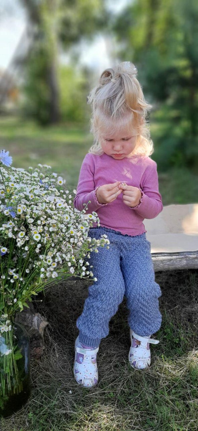 May include: A young child wearing a pink long-sleeved shirt and blue knitted trousers is sitting on a wooden surface. The child is holding a small white flower in their hand. The child is wearing white sandals with pink accents.