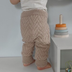 May include: A baby wearing a pair of beige knitted trousers with a chevron pattern. The trousers have a ribbed waistband and cuffs.