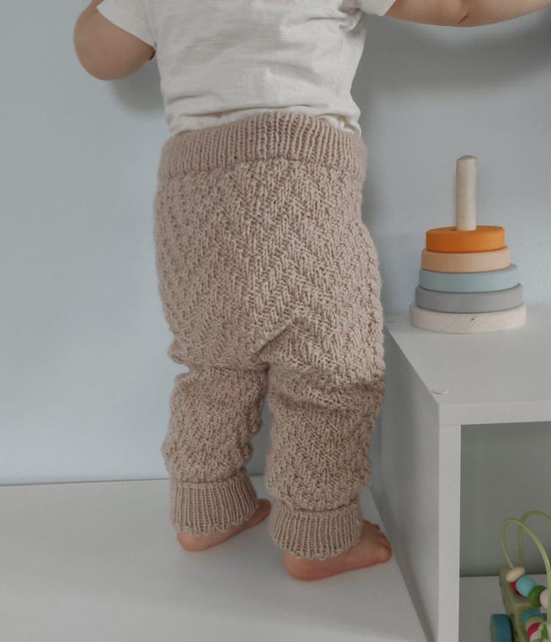 May include: A baby wearing a pair of beige knitted trousers with a chevron pattern. The trousers have a ribbed waistband and cuffs.