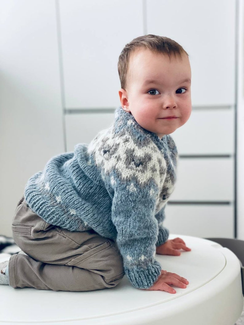 May include: A young child wearing a blue and white patterned sweater and brown pants is kneeling on a white surface. The child is looking at the camera.