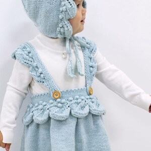 May include: A light blue knitted baby dress with a matching bonnet. The dress has a scalloped hem and straps with wooden buttons. The bonnet has a scalloped edge and a tie.