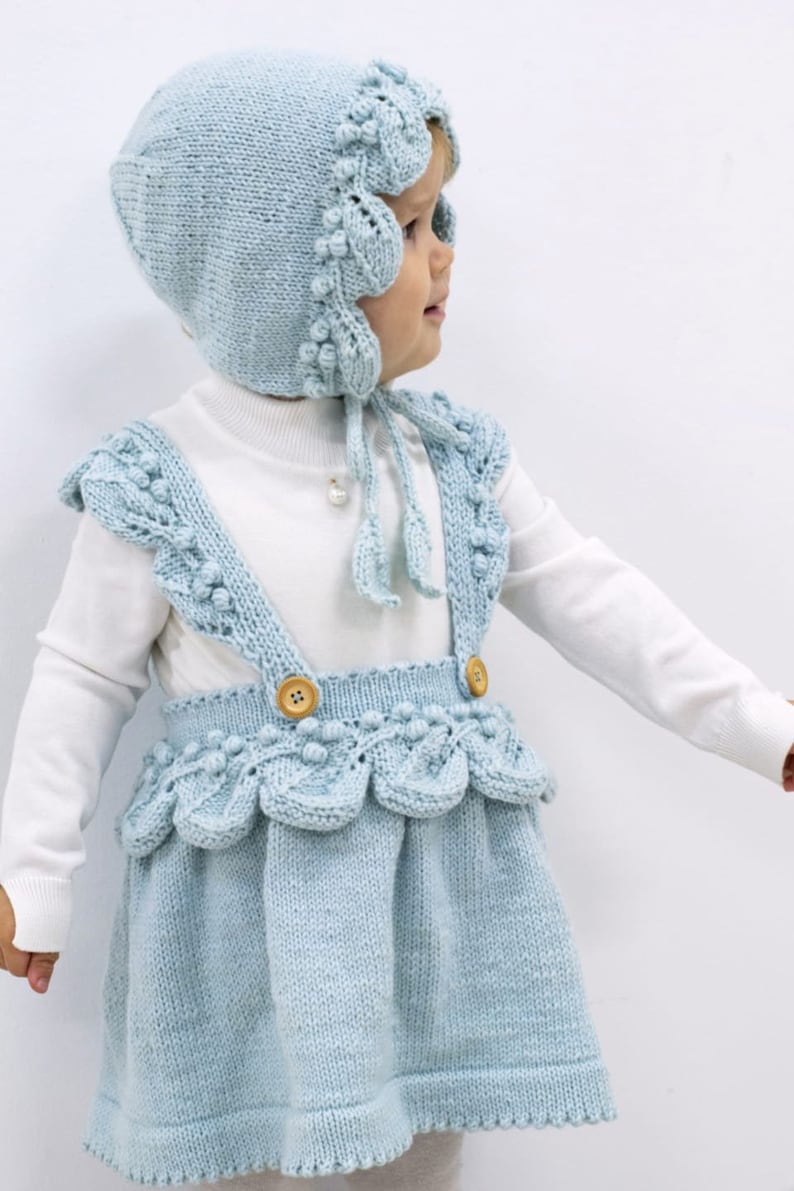 May include: A light blue knitted baby dress with a matching bonnet. The dress has a scalloped hem and straps with wooden buttons. The bonnet has a scalloped edge and a tie.