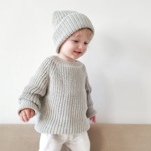 Puede incluir: Un ni&ntilde;o peque&ntilde;o con un su&eacute;ter de punto gris claro y una boina gris claro. El ni&ntilde;o est&aacute; de pie frente a una pared beige claro.