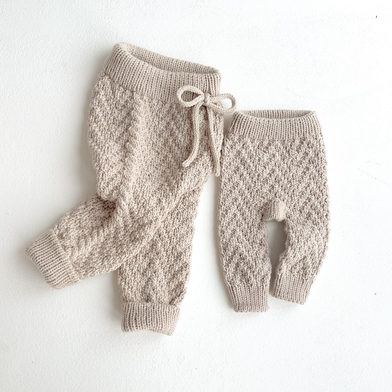 May include: A set of two knitted trousers in a light beige colour. The trousers are made from a soft, textured fabric and have a relaxed fit. The larger trousers have a drawstring waist.