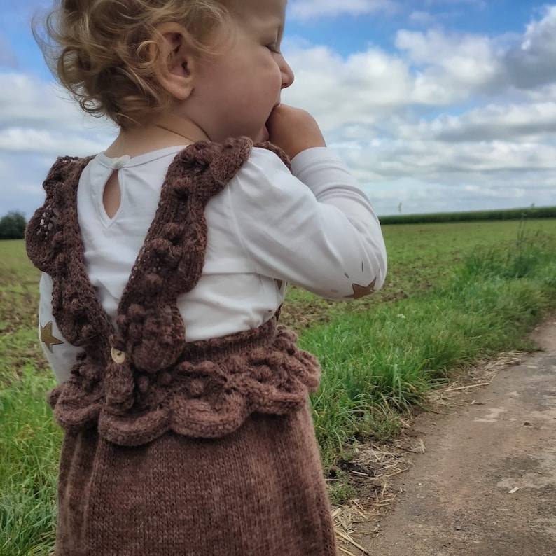 May include: A brown knitted baby overall with a white shirt underneath. The overall has a scalloped hem and a decorative button on the front. The baby is wearing the overall and looking off to the side.