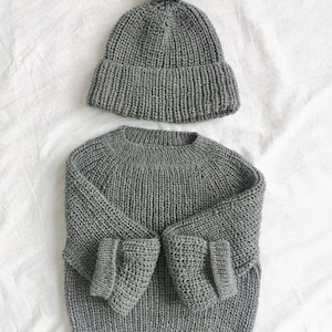 Puede incluir: Un su&eacute;ter de punto gris y un gorro a juego con un pomp&oacute;n en la parte superior.