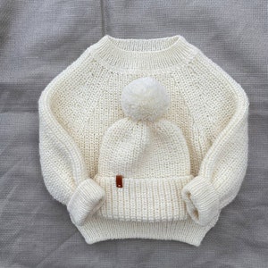 Puede incluir: Un su&eacute;ter de punto blanco y un gorro a juego con un pomp&oacute;n blanco.