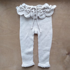 Op de afbeelding: Witte gebreide babybroek met een gekrulde rand rond de taille. De rand heeft een strik en witte pompoentjes.