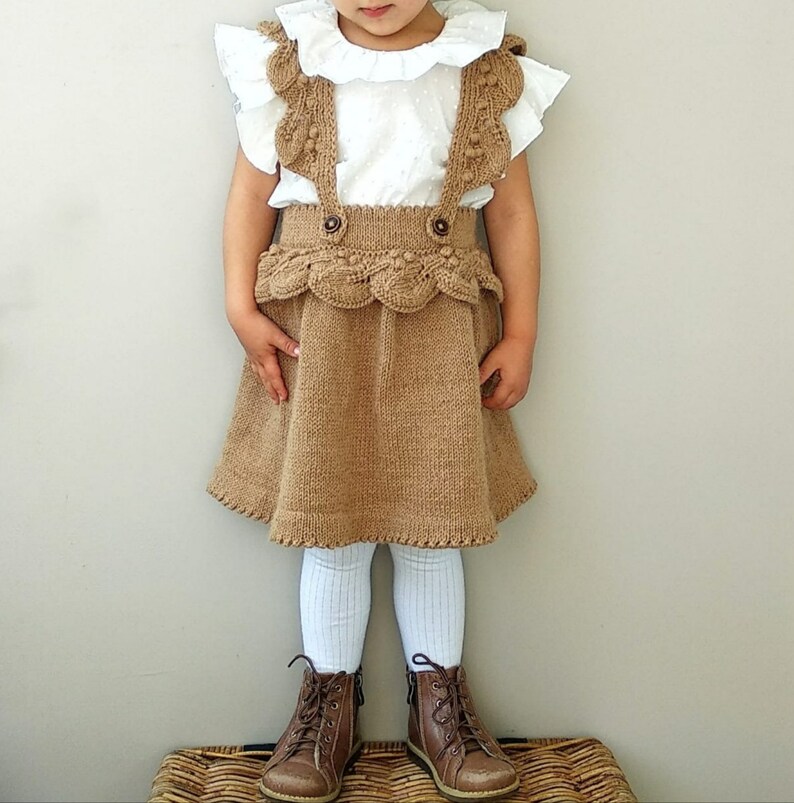May include: A brown knitted skirt with a scalloped hem and a white ruffled blouse. The skirt has a decorative strap detail and the blouse has a white collar.