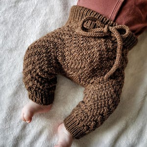 May include: Brown knitted baby trousers with a drawstring waist. The trousers are made with a chevron pattern.