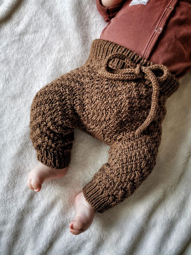 May include: Brown knitted baby trousers with a drawstring waist. The trousers are made with a chevron pattern.