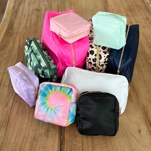 May include: An assortment of small zippered pouches in various colours and patterns, including pink, white, green, blue, and tie-dye. The pouches are made of fabric and have gold zippers.