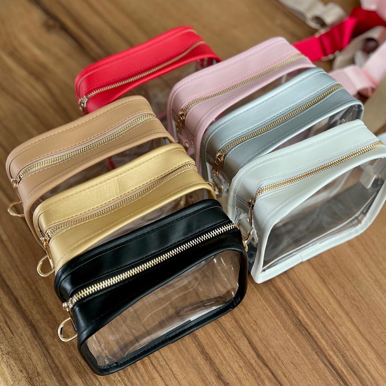 May include: Six clear plastic bags with zipper closures and a leather flap. The bags are in different colors: red, pink, gray, white, gold, and black.