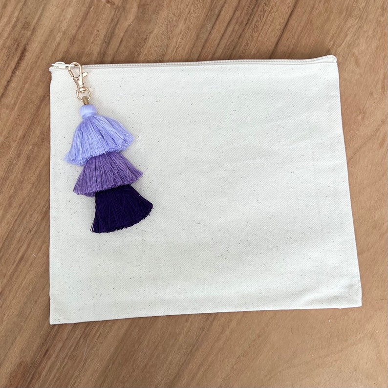 May include: A white canvas zippered pouch with a purple, blue, and lavender tassel attached to the zipper pull.