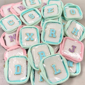 May include: A collection of small, clear, zippered pouches with a variety of letters on the front. The pouches are in pink and light blue colours. The letters are made of a fluffy material and are in a variety of colours.