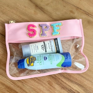May include: A pink cosmetic bag with the letters "SPF" in colorful, textured patches. The clear front reveals sunscreen products, including a tube of Neutrogena Ultra Sheer and a Banana Boat Ultra Defense spray.
