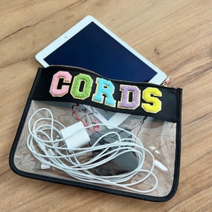 May include: A clear plastic zippered pouch with a black fabric flap. The flap has the word "CORDS" embroidered in colorful letters. The pouch contains a white charging cable and a black phone charger.