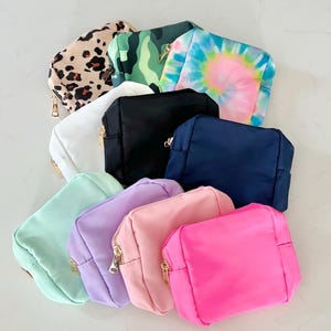 May include: A selection of small, rectangular cosmetic bags in various colours and patterns. The bags include solid colours like white, black, navy, pink, and mint green, as well as patterns like tie-dye, camouflage, and leopard print. Each bag has a gold zip.