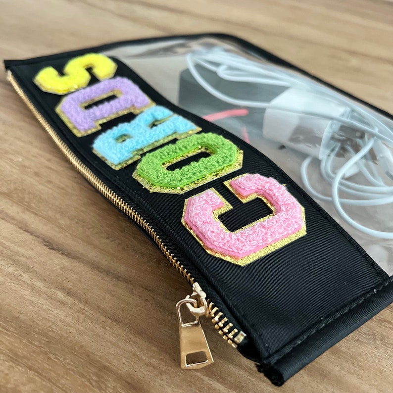 以下が含まれることがあります： カラフルで質感のある文字で「SABGC」と書かれた黒いジッパーポーチ。ポーチは金色のジッパーと透明な上部があり、内部の電子アクセサリーが見えます。