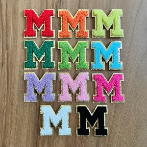 Op de afbeelding: Negen vilt letter patches in verschillende kleuren, elk met een gouden omtrek. De letters spellen het woord "MMMMMMMM" uit.