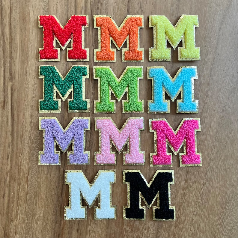 Op de afbeelding: Negen vilt letter patches in verschillende kleuren, elk met een gouden omtrek. De letters spellen het woord "MMMMMMMM" uit.
