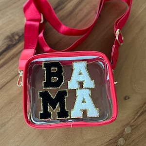 May include: A clear plastic crossbody bag with a red frame and a gold chain strap. The bag has the letters "BAMA" in black and white glitter on the front.