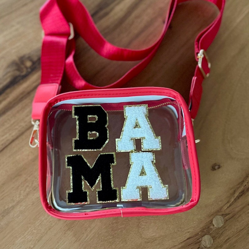 May include: A clear plastic crossbody bag with a red frame and a gold chain strap. The bag has the letters "BAMA" in black and white glitter on the front.