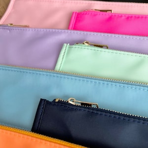 Pode incluir: Um conjunto de cinco estojos com z&iacute;per em v&aacute;rias cores pastel, incluindo rosa, lavanda, verde claro, azul claro e azul marinho. Os estojos s&atilde;o feitos de um tecido macio semelhante ao nylon e t&ecirc;m z&iacute;peres dourados.