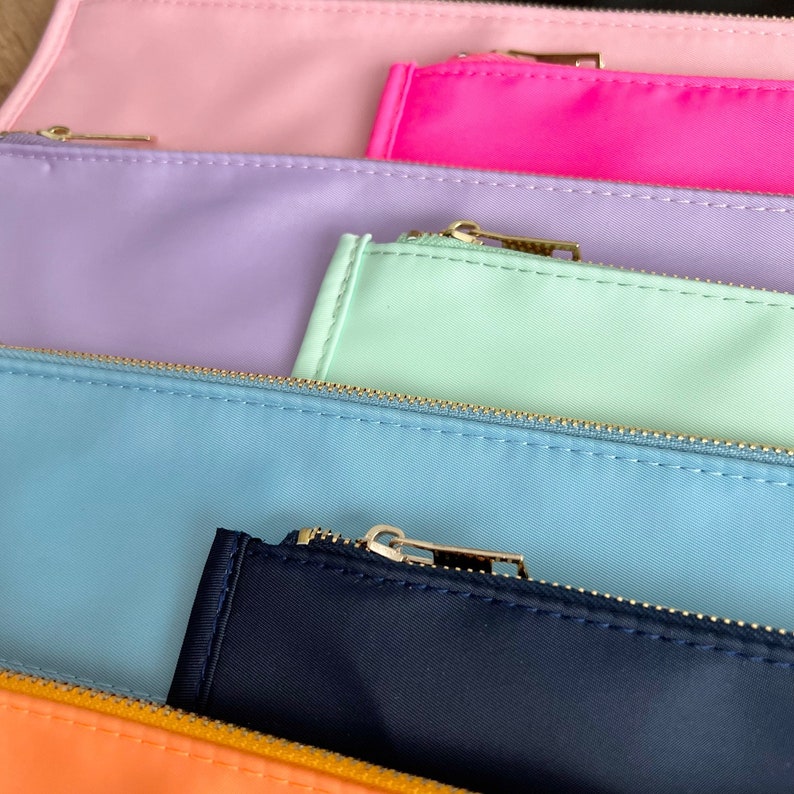 Pode incluir: Um conjunto de cinco estojos com z&iacute;per em v&aacute;rias cores pastel, incluindo rosa, lavanda, verde claro, azul claro e azul marinho. Os estojos s&atilde;o feitos de um tecido macio semelhante ao nylon e t&ecirc;m z&iacute;peres dourados.