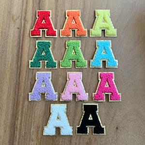 Op de afbeelding: Tien vilt letter patches in verschillende kleuren, elk met de letter 'A'. De patches zijn gerangschikt in twee rijen van vijf, met kleuren die afwisselen van rood naar oranje, geel, groen, blauw, paars, roze, wit en zwart.
