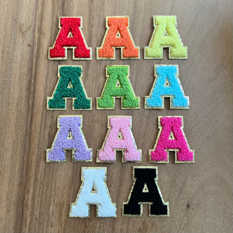 Op de afbeelding: Tien vilt letter patches in verschillende kleuren, elk met de letter 'A'. De patches zijn gerangschikt in twee rijen van vijf, met kleuren die afwisselen van rood naar oranje, geel, groen, blauw, paars, roze, wit en zwart.