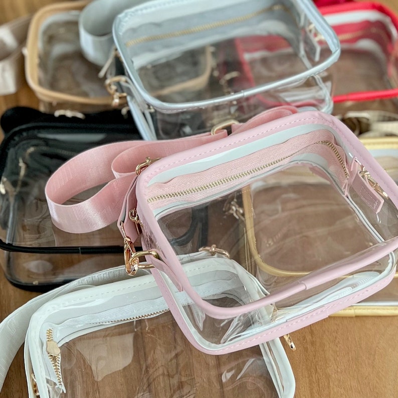 May include: A stack of clear plastic bags with pink, white, and gray trim. The bags have gold zippers and straps.