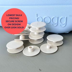 May include: A close-up of white plastic screw-on buttons for a pair of Bogg shoes. The buttons are scattered on a grey surface. The text "LOWEST BULK PRICING! SECURE SCREW ON DESIGN! OVER 100K SOLD!" is visible in red on a pink circle in the background.