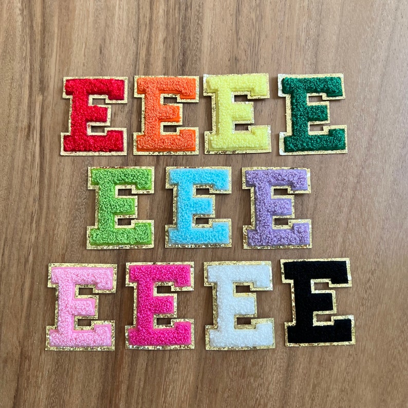 Op de afbeelding: Negen strijkbare patches in verschillende kleuren, elk met een pluche, witte letter "E" met een gouden rand. De kleuren omvatten rood, oranje, geel, groen, lichtgroen, lichtblauw, lavendel, roze, wit en zwart.