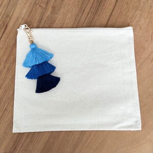 May include: A white canvas zippered pouch with a blue, teal, and navy tassel attached to the zipper pull.