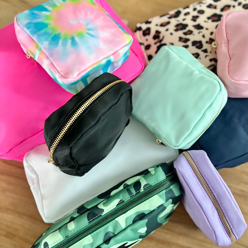 Op de afbeelding: Een verzameling van negen kleine ritszakjes in verschillende kleuren en patronen, waaronder tie-dye, camouflage, effen zwart, effen wit, effen lichtblauw, effen lavendel en luipaardprint.
